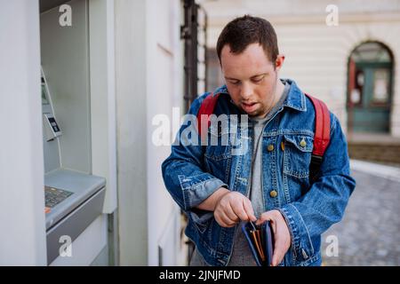 Happy young man with Down sydrome using a street ATM machine and withdrawing money. Stock Photo