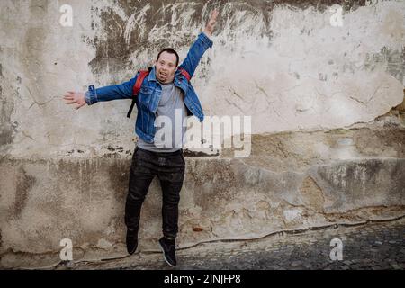 Cheerful young man with Down sydrome jumping in the street, in front of wall with raising arm, happy gesture. Stock Photo