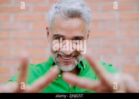 Mature man with happy expression, showing peace sign with hand and looking at camera, on street background. Stock Photo