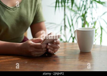 Close-up shot of unrecognizable woman wearing T-shirt sitting at cafe table with cup of fragrant coffee and texting with friend on smartphone, blurred background Stock Photo