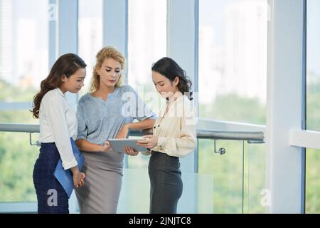 Pretty female business executives discussing information on tablet compuer in office corridor Stock Photo