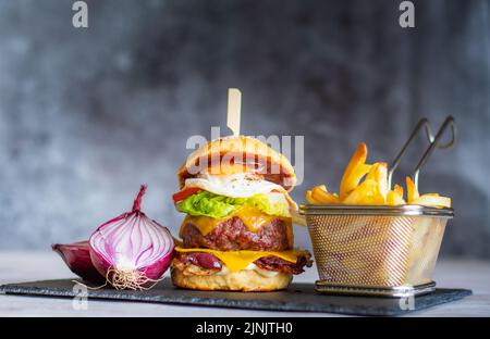 Tasty homemade cheeseburger with french fries placed on black desk, gray background. Cheese, bacon, egg, salad, tomato and onion in light bread. Unhea Stock Photo