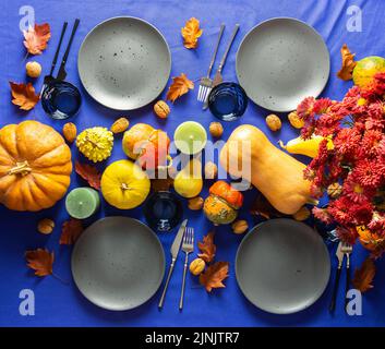 Thanksgiving table setting, bouquet with dry branches, various pumpkins and candles, plates with cutlery and glasses on a blue-purple tablecloth Stock Photo