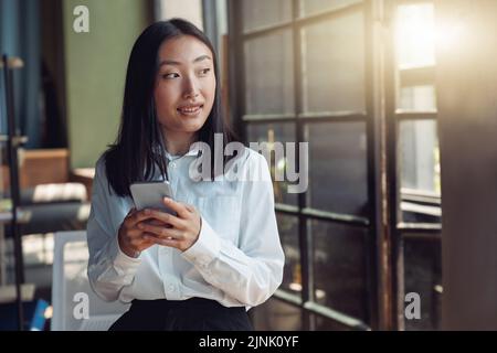 Asian businesswoman standing at office with phone and looking at window Stock Photo