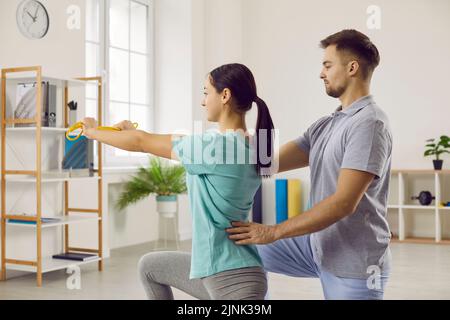 Young female patient with elastic band does stretching exercise with male physiotherapist. Stock Photo