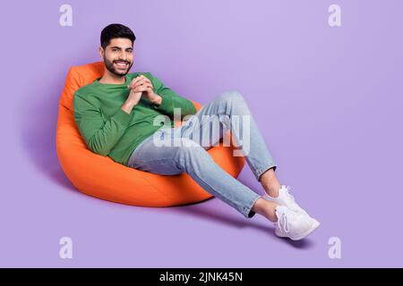 Full size photo of young latin man sit comfy beanbag hold hands toothy smile wear stylish green clothes isolated on purple color background Stock Photo