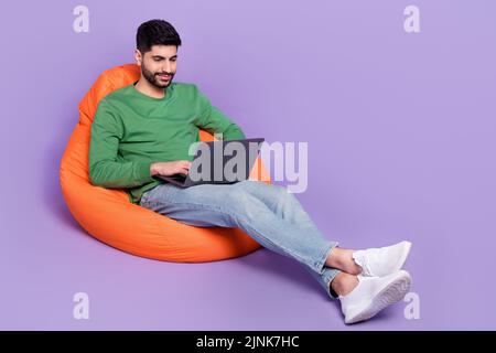 Full body photo of young latin guy sitting coding new software entrepreneur wear stylish green garment isolated on purple color background Stock Photo