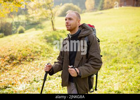 Sporty tourist man with trekking poles and a backpack travels through the mountain hills. Smiling man enjoys the beauty of nature. Concept of hiking, Stock Photo