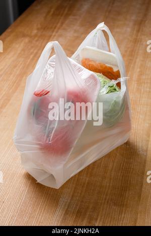 Close up of plastic bag with various vegetables inside which are wrapped in cling film. Grocery package from supermarket or grocery store stands on wo Stock Photo