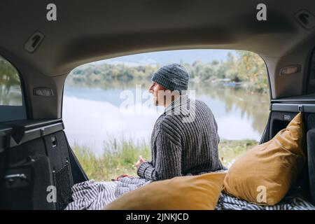 Midle-aged smiling man dressed in warm knitted clothes sitting in the opened Car trunk and enjoying a beautiful autumnal mountain lake view after nigh Stock Photo