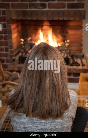 Woman from behind sitting on the floor in front of the comfortable warmth of a fireplace. Stock Photo