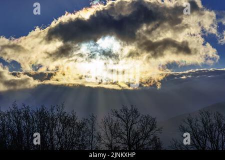 Sunrise with the sun through the clouds and sunbeams coming out of the snowy mountains. Stock Photo
