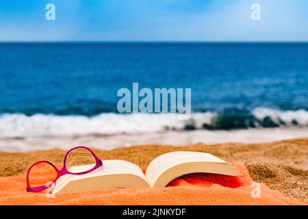 book and glasses on the beach towel with the sea in the background Stock Photo