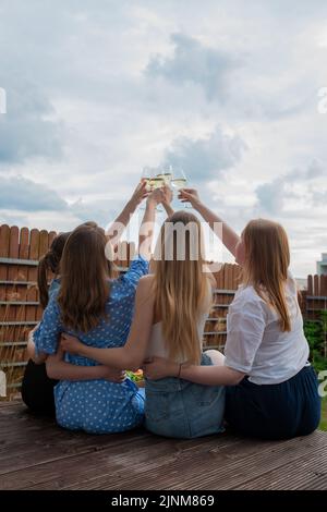 Back view of group of young women with long hair raising clinking glasses with white wine champagne on sky background. Stock Photo