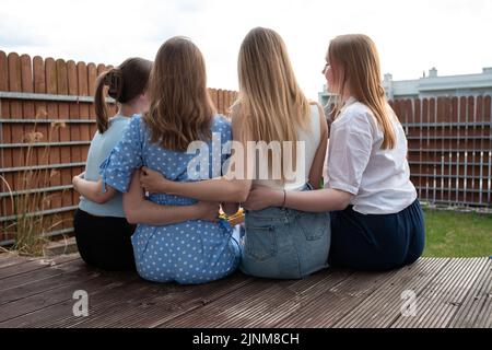 Back view of group of young women with long hair sitting on wooden veranda, embracing hugging in backyard. Summer. Stock Photo