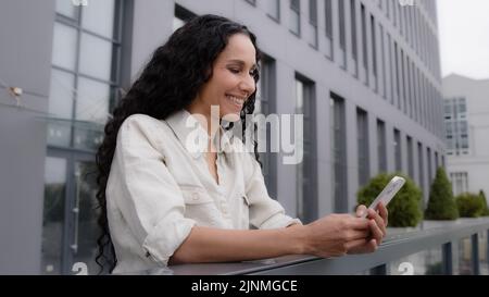 Side view Caucasian Hispanic 30s happy smiling woman standing on balcony terrace in city female businesswoman with phone ecommerce shopping online Stock Photo