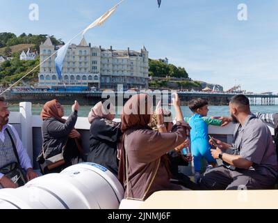 Llandudno, Clwyd, Wales, August 07 2022: People enjoying a boat trip and taking photos with the Grand Hotel and pier in the background. Stock Photo