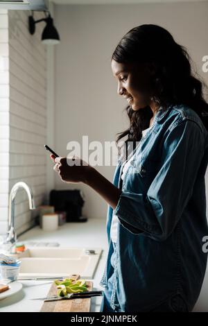 Black young woman using mobile phone while cooking at home Stock Photo