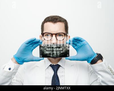 Male dentist cover his mouth with a smartphone with X-ray of his teeth on a screen of device. Stock Photo