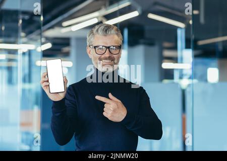Senior successful gray-haired businessman in office, showing phone screen to camera with white screen background, man pointing with finger on copy space Stock Photo