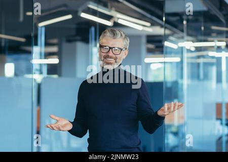 Senior gray-haired male businessman looking into web camera and smiling standing in office online meeting, teachers cheerfully waving hands, video call remote conversation colleagues web cam view Stock Photo