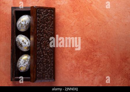 carved wooden treasure box with 3 golden eggs inside on a textured orange background Stock Photo