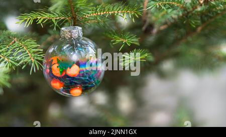 Green Christmas tree with beautiful round glass decoration on a white blurry background. Closeup of Xmas ornament hanging on a natural spruce branch. Stock Photo