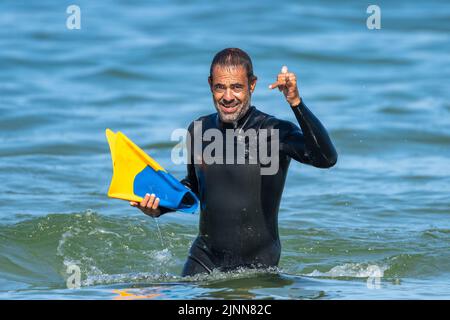 Middle-aged dark-haired man with sea water up to his waist dressed in a thermal suit, with fins in one hand and greeting with the other. Stock Photo