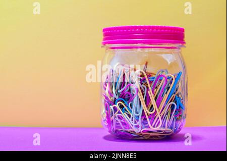 Colorful paperclips in a plain jar and sealed lid with yellow background and copy space, creative concept. Stock Photo