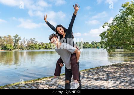 Happy couple. Handsome young man carrying his beautiful girlfriend on his back. She is raising her hands and looking at camera Stock Photo