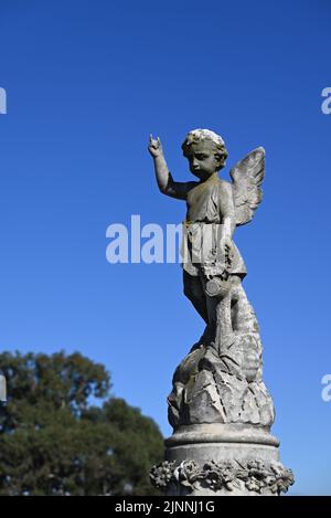 Stone sculpture of a child angel, or cherub, pointing skyward whilst looking down, atop a grave during a clear day Stock Photo