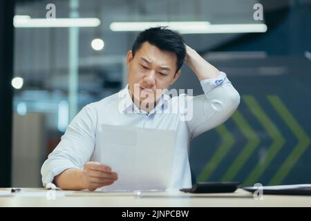 Stressed businessman holding face with his hands because having trouble. Bankrupt Asian Male is sitting with laptop computer and paperwork on desk at workplace in office. Business Mistake Financial. Stock Photo