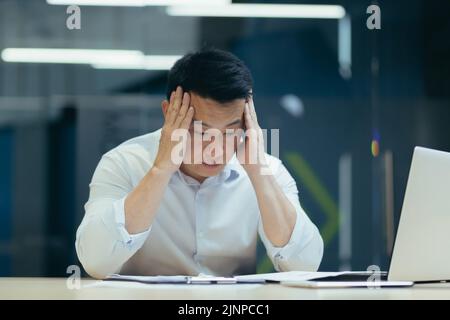 Stressed businessman holding face with his hands because having trouble. Bankrupt Asian Male is sitting with laptop computer and paperwork on desk at workplace in office. Business Mistake Financial. Stock Photo