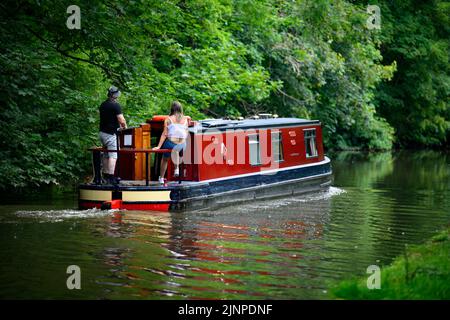 Couple relaxing on red cruiser-style narrow boat moving slowly along scenic quiet rural Leeds Liverpool Canal - Bingley, West Yorkshire, England, UK. Stock Photo