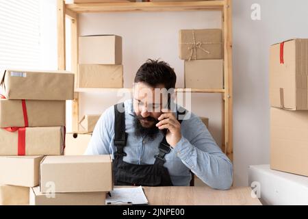 bearded man online seller confirming orders from customer on the phone. E-commerce male business owner using phone in store warehouse with boxes for Stock Photo