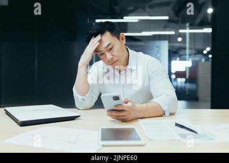 Confused Asian guy holding smart phone feels concerned thinking over received message. Mobile phone everyday usage, unpleasant news, waiting for important call, low signal device problems concept Stock Photo