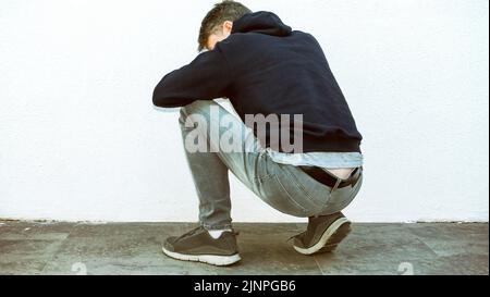 An image of a young man in a black hoodie, blue jeans, and gray sneakers squatting with his head down, evoking a contemplative mood. Stock Photo