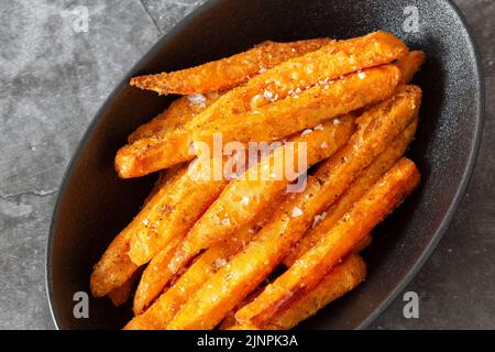 Sweet potato fries, seasoned with sea salt, in a black dish bowl.  On a dark stone background Stock Photo
