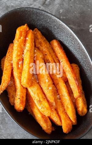 Sweet potato fries, seasoned with sea salt, in a black dish bowl.  On a dark stone background Stock Photo
