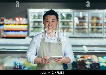 portrait of handsome asian staff man salesman in apron standing and looking at camera in grocery store supermarket. Male worker seller or small business owner. indoor. food, groceries market smile Stock Photo