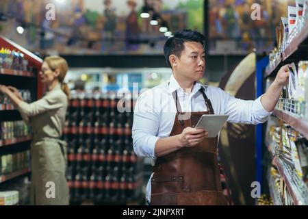 Two workers in a supermarket in the grocery department arrange products, an Asian man with a tablet computer and a woman in aprons among the products on the shelves and racks Stock Photo