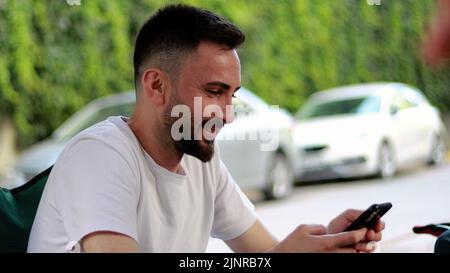 Bearded and handsome young man using mobile phone outdoors. Happy smiling man in white t-shirt looking at mobile phone screen and reading messages Stock Photo