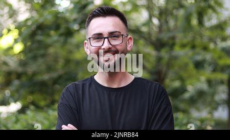 Portrait of young handsome man with beard and glasses. Young man in black shirt smiling looking at camera. Outdoor nature background Stock Photo