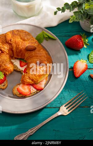 Appetizing French croissant with strawberries, almonds and fragrant mint leaves on a plate on a turquoise background. Continental breakfast for one pe Stock Photo