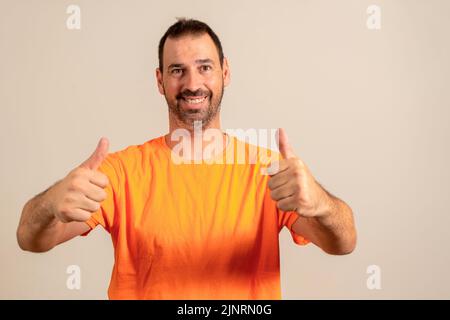 Handsome but unkempt, unshaven and out of shape man poses with thumbs up while smiling at camera, isolated over beige background. Stock Photo