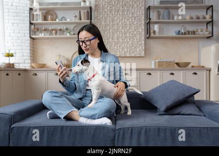 Young beautiful asian woman sitting on sofa at home and using phone happily, teenager with pet jack russell dog having fun together Stock Photo
