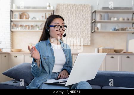 Young beautiful asian woman studying remotely sitting in living room on sofa, female student using headset and laptop for online distance learning and video call, teenager wearing glasses and shirt at home Stock Photo
