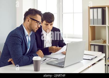 Two serious young men sitting at an office desk and looking at their laptop computer together Stock Photo