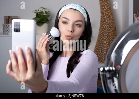 influencer, beauty blogger, content creator or vlogger making a selfie on a mobile phone front camera. attractive Young woman holds a cosmetic brush f Stock Photo