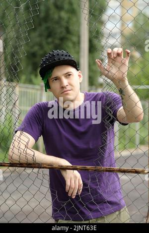 Young man with green dreadlocks in black cap, purple t-shirt posing through hole of lattice fence Stock Photo